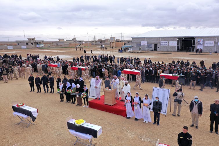 Two caskets sit on stands as people stand formally in a row behind them, in a sandy landscape with a few low buildings in the background.