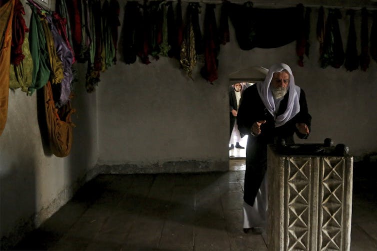 A man with a white covering around his head holds his hands in prayer inside a darkened room with colorful pieces of fabric on the wall.