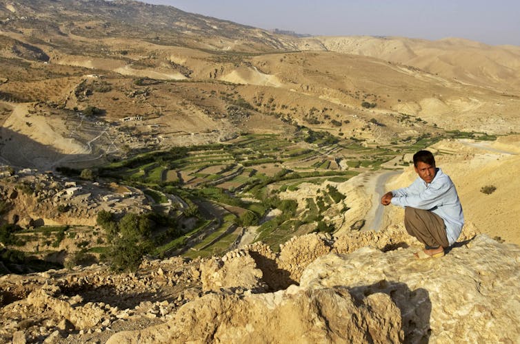A man crouches on a plateau overlooking a sweeping, dry landscape of hills.