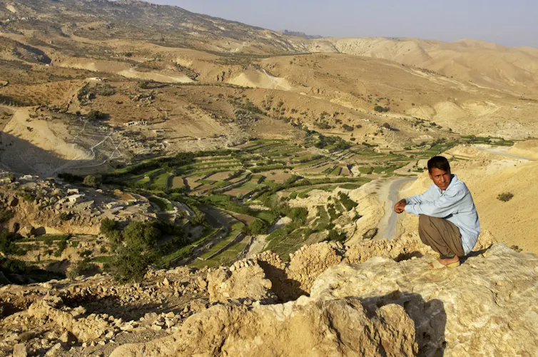 A man crouches on a plateau overlooking a sweeping, dry landscape of hills.