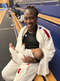 A Black woman in a judo uniform, which consists of a white tunic, pants, and a belt, breastfeeds an infant while sitting on a bench in a gymnasium