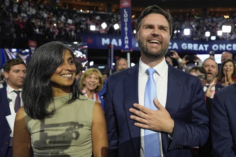 A bearded white man in a blue suit and light blue tie stands next to a woman in a green dress. Both are smiling.