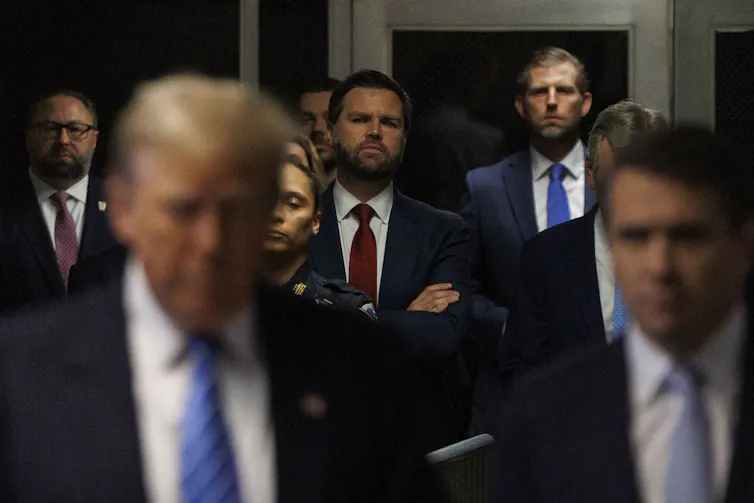 A man in a beard with his arms folded is in the background while Trump is out of focus in the foreground.