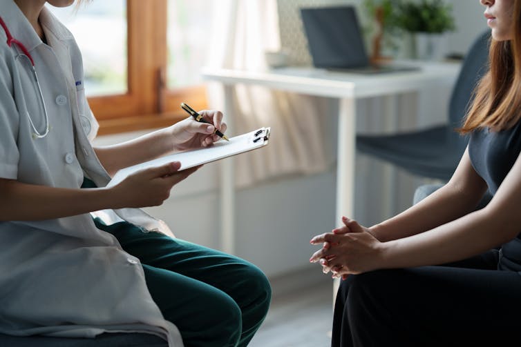 A doctor writes on a clipboard while seated in front of a young woman. They are both only seen from the neck down.