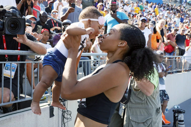 A Black woman in a cropped tank top lifts a baby up, who is wearing a headband with a flower on it, while standing in front of a set of bleachers full of people