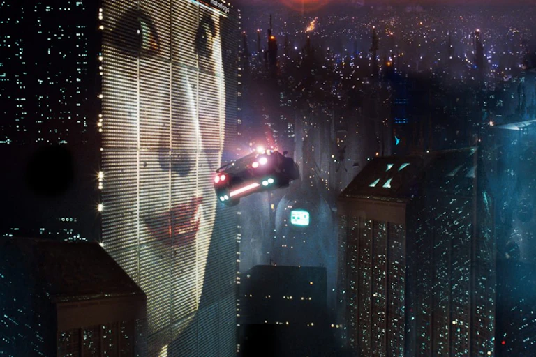 cena do filme Blade Runner, com um carro voador passando por uma tela gigante na lateral de um edifício