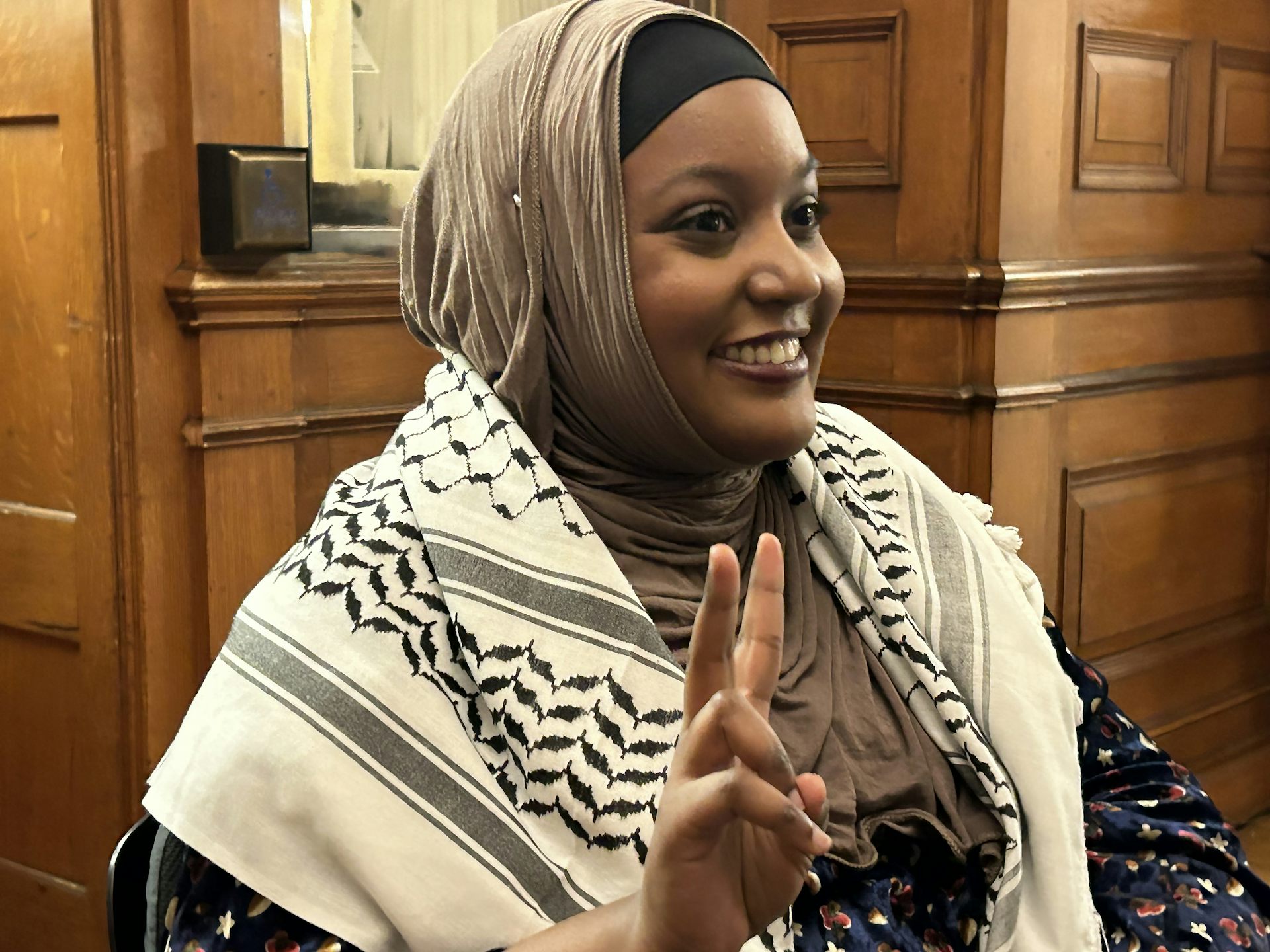 A Black woman in a hijab wearing a keffiyeh on her shoulders makes a victory sign with her hand