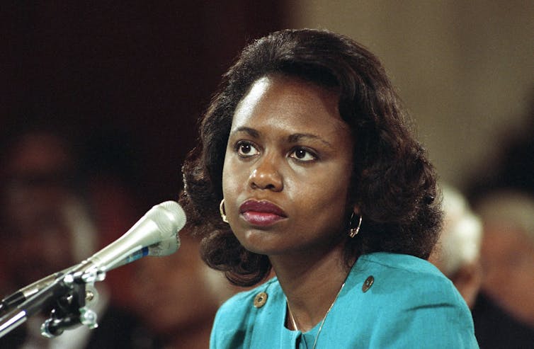 A Black woman sits at a table in front on a microphone