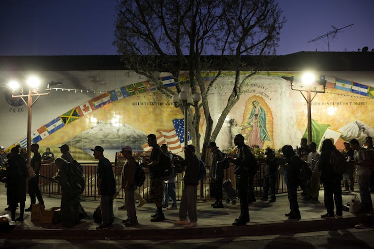 A line of people wait in front of a mural outdoors, faintly illuminated under streetlights.