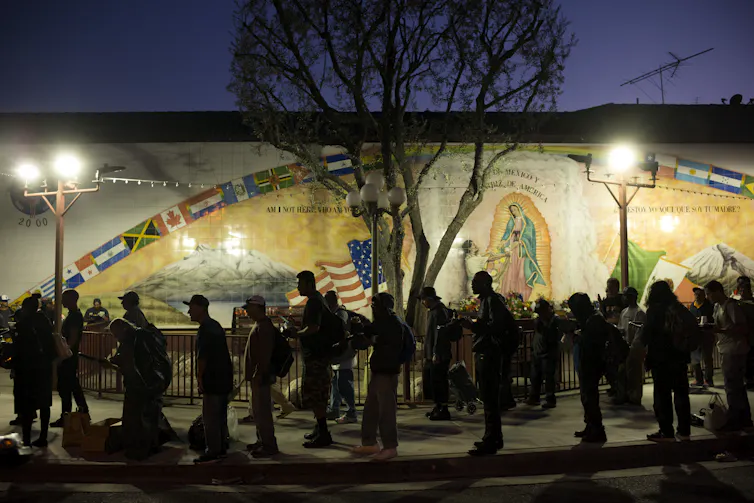 A line of people wait in front of a mural outdoors, faintly illuminated under streetlights.