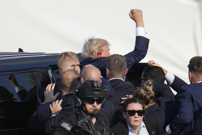 Donald Trump climbs into a car while waving and being surrounded by security staff