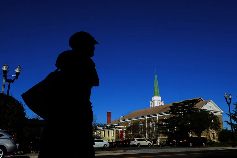 A person in a hat and coat, silhouetted against a dark blue sky, walks across the street from a church.