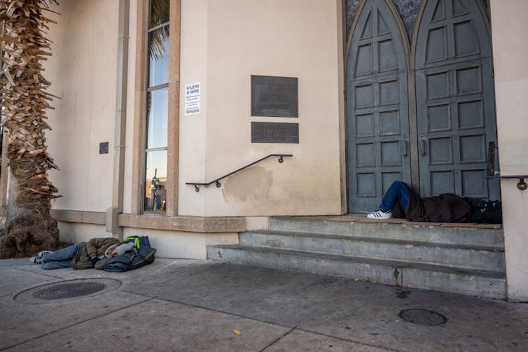 Two people sleep on the concrete sidewalk and stairs outside a building with carved wooden doors and a tall window.