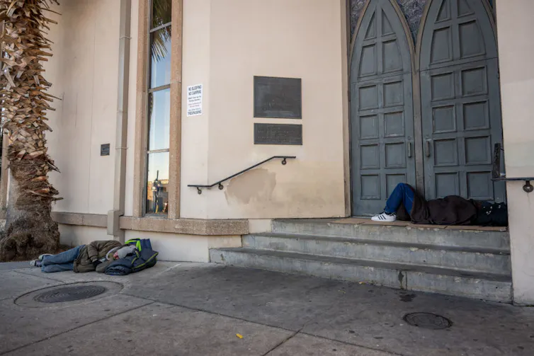 Two people sleep on the concrete sidewalk and stairs outside a building with carved wooden doors and a tall window.