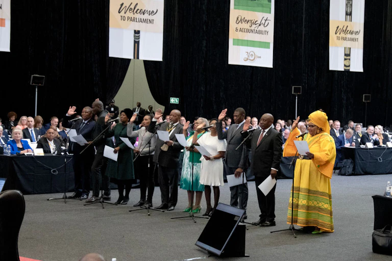 A row of people hold up their right hands, holding a piece of paper in their other hand, with microphones in front of them. Other people are seated in the background and posters on the wall say 'welcome to parliament'.