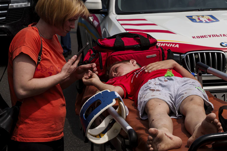 A boy in a red T-shirt and shorts lies on a gurney as a rescuer attends to him.
