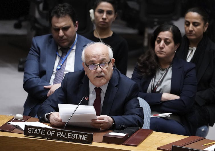 An older man with white hair and glasses wears a dark suit and speaks into a microphone while seated, with a placard that says 'State of Palestine' in front of him.