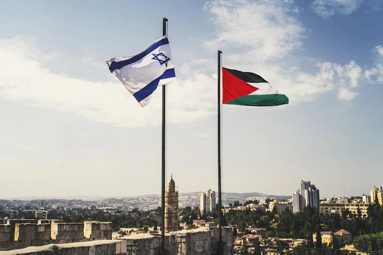 An Israeli flag is raised next to a Palestinian flag, against a blue sky.
