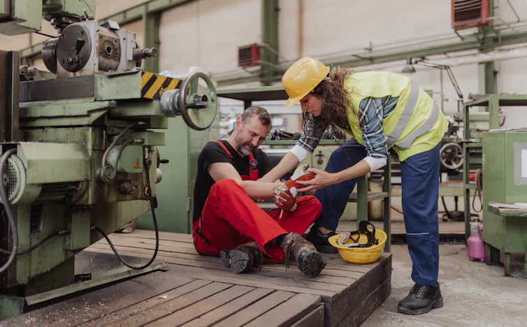 A woman in a construction hat and vest holds the hand of a man in red overalls, who is sitting on the floor beside heavy machinery with a pained look on his face