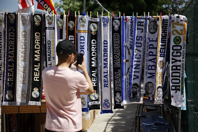 A man takes a photo of scarves with soccer team names printed on them hanging from a clothes line