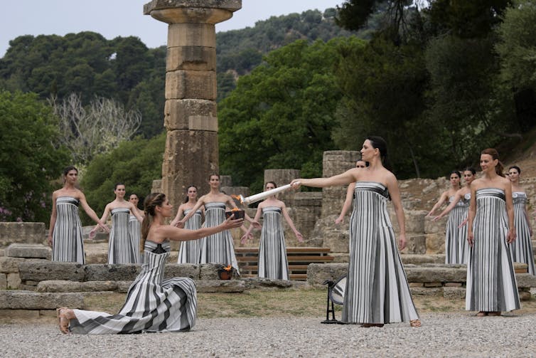 A group of women wearing dresses stylized to look like Greek columns stand among ancient ruins. One of the women kneels and holds a bowl in her outstretched arms while another lights the bowl with a torch
