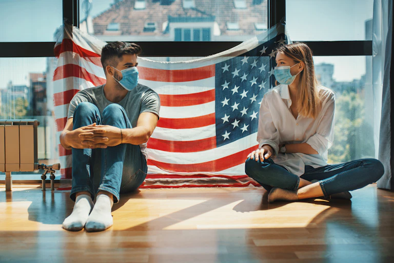 A seated, masked man and woman with their backs to an American flag hanging on a window