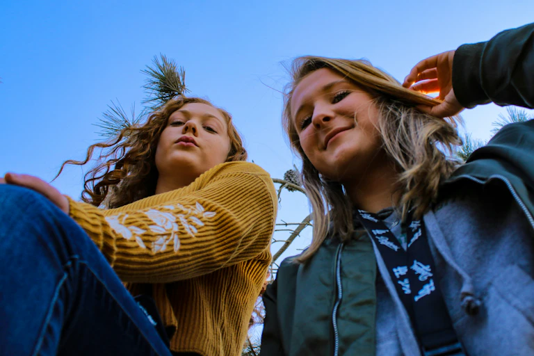 Two young women stand sit by side, outdoors, with blue sky and a plant behind them.