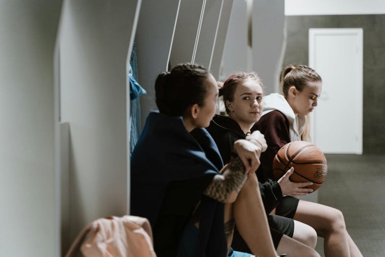 Three women sit in a locker room in sports clothes. One holds a basketball.