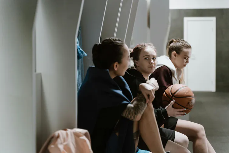 Three women sit in a locker room in sports clothes. One holds a basketball.