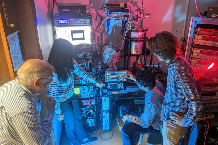A group of people in a blue and red lit room clustered around complex looking machinery.