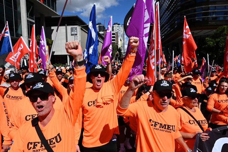 A group of men in bright orange shirts raise their fists and yell at a rally