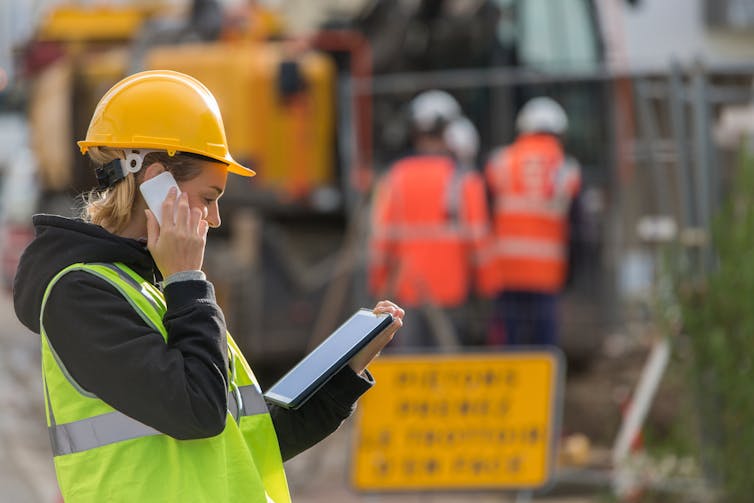 A woman in a high-vis vest and a hard hat makes a phone call on a construction site.