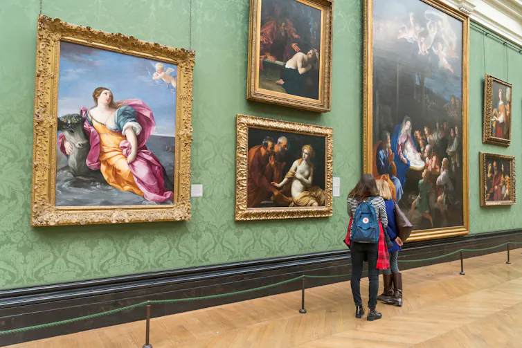 Two people look at a gallery wall of paintings.