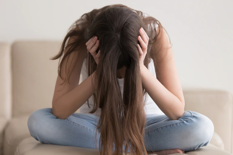 Teenage girl with long hair, sitting cross-legged on sofa, head in hands, looking down