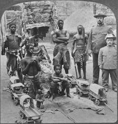 A vintage photo with people posing in a rural village setting as if in a human zoo. Six are Africans in animal cloths and two are Europeans in military uniforms, one a short person.
