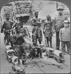 A vintage photo with people posing in a rural village setting as if in a human zoo. Six are Africans in animal cloths and two are Europeans in military uniforms, one a short person.