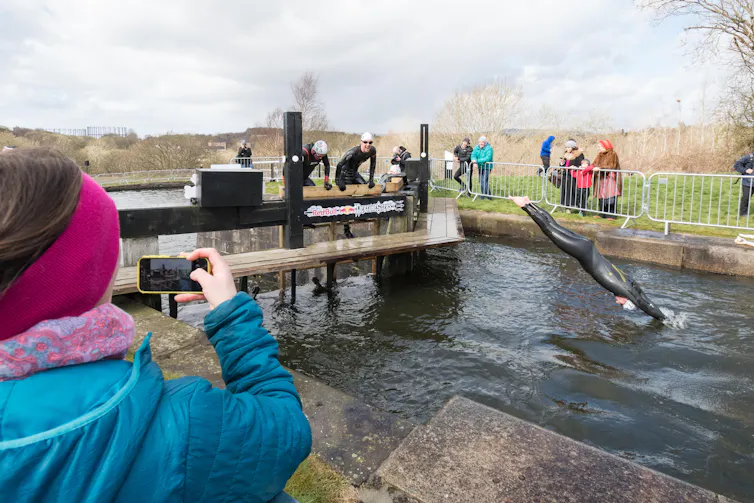 Man in wetsuit dives from lock into canal, people watching