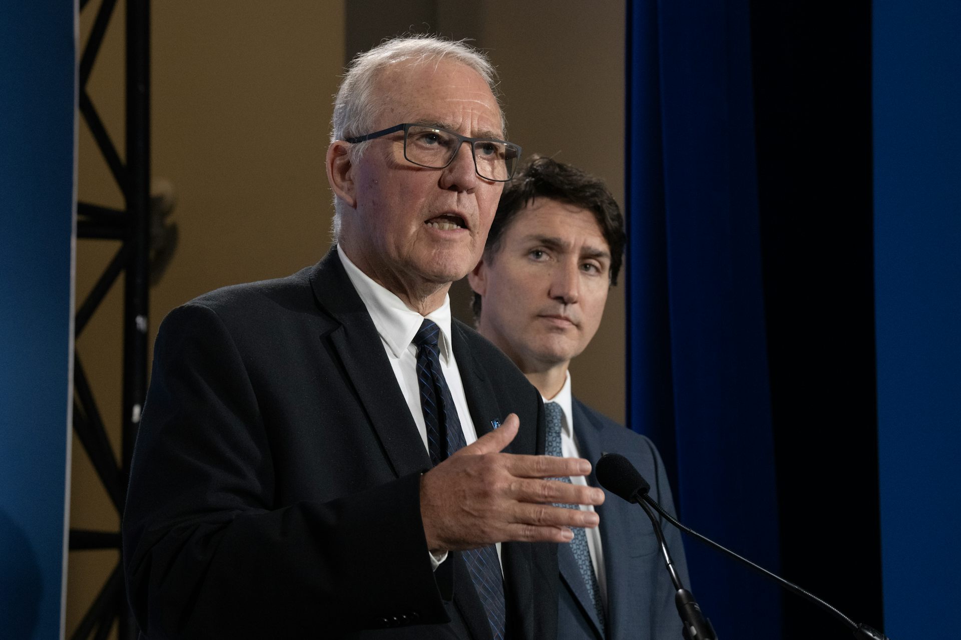 A man with thinning grey hair and glasses speaks into a microphone as a dark-haired younger man look on.