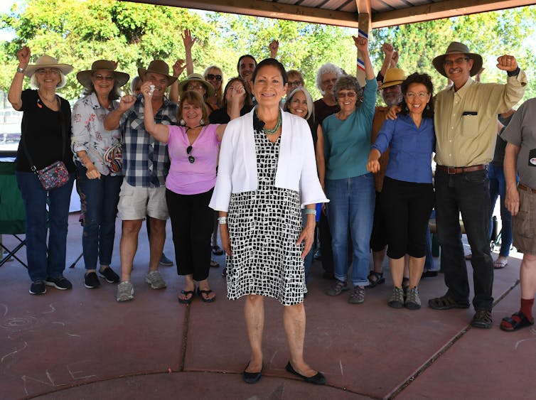 Deb Haaland stands smiling in front of a small cheering crowd of Native Americans, many wearing cowboy hats
