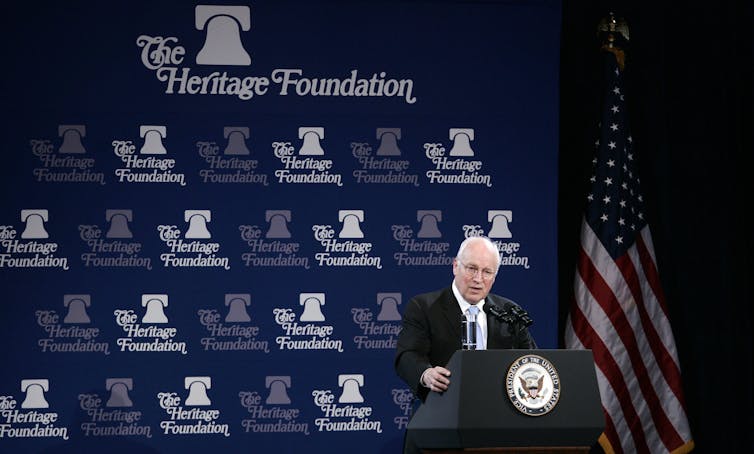 An older white man stands at a podium and in front of a blue backdrop that says 'The Heritage Foundation'
