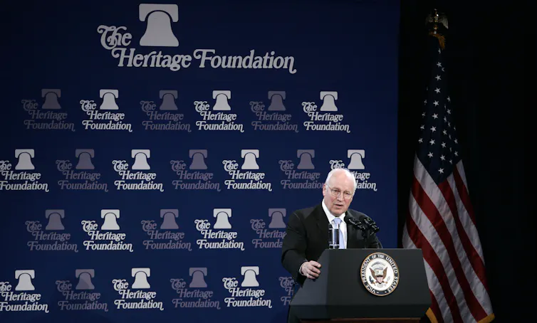 An older white man stands at a podium and in front of a blue backdrop that says 'The Heritage Foundation'