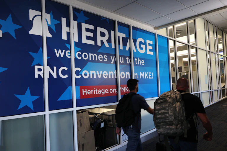 Two people walk past a large blue sign that says in white writing, 'Heritage welcomes you to the RNC in MKE!'