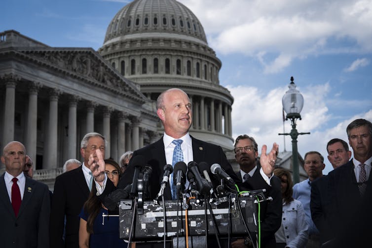 A white man stands at a podium with several microphones, in front of other people in suits and in front of the Capitol building.