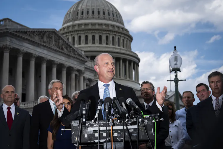 A white man stands at a podium with several microphones, in front of other people in suits and in front of the Capitol building.