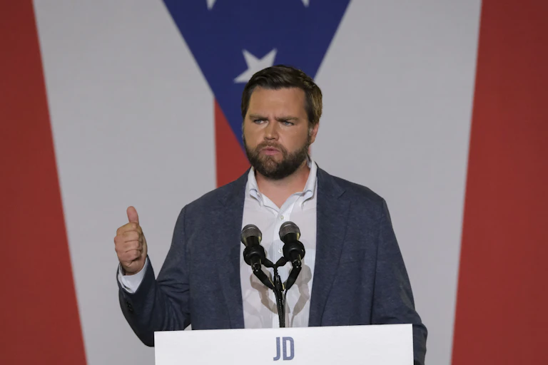 A bearded man speaking from the lectern with the American flag in the background.