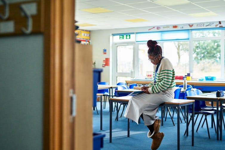 A teacher sits on a desk in an empty classroom.