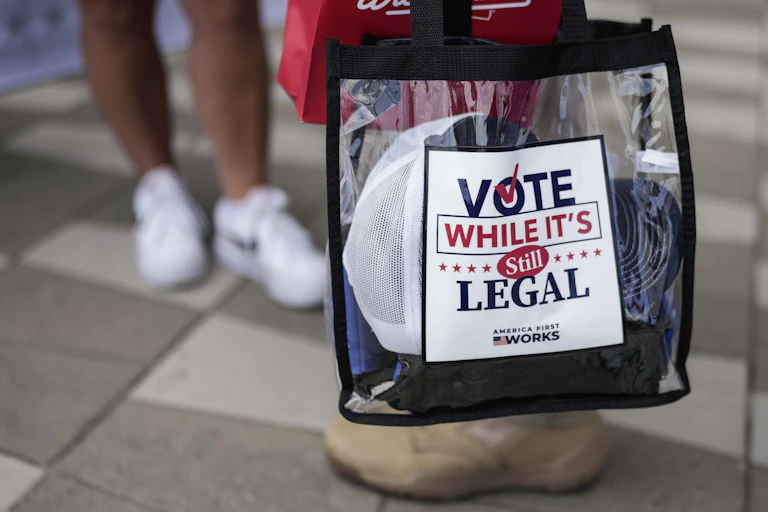 A clear plastic tote bag with a sign on it that says 'VOTE WHILE IT'S STILL LEGAL'