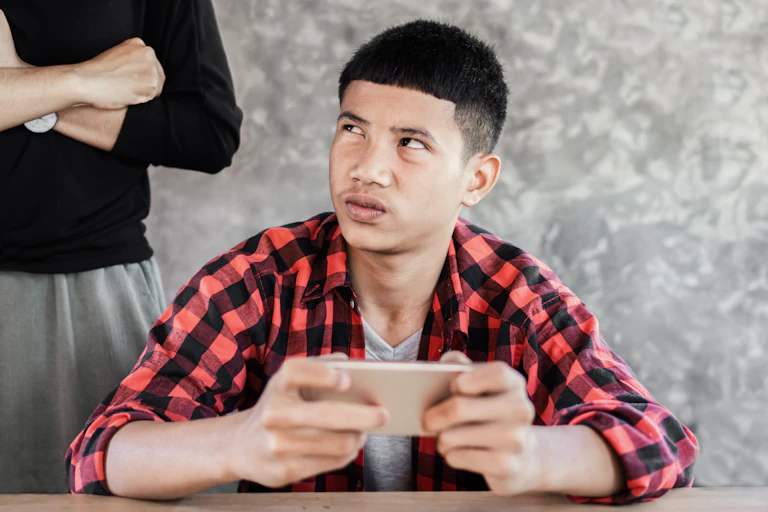 a boy holds an iphone and looks up at an adult standing behind him with their arms crossed