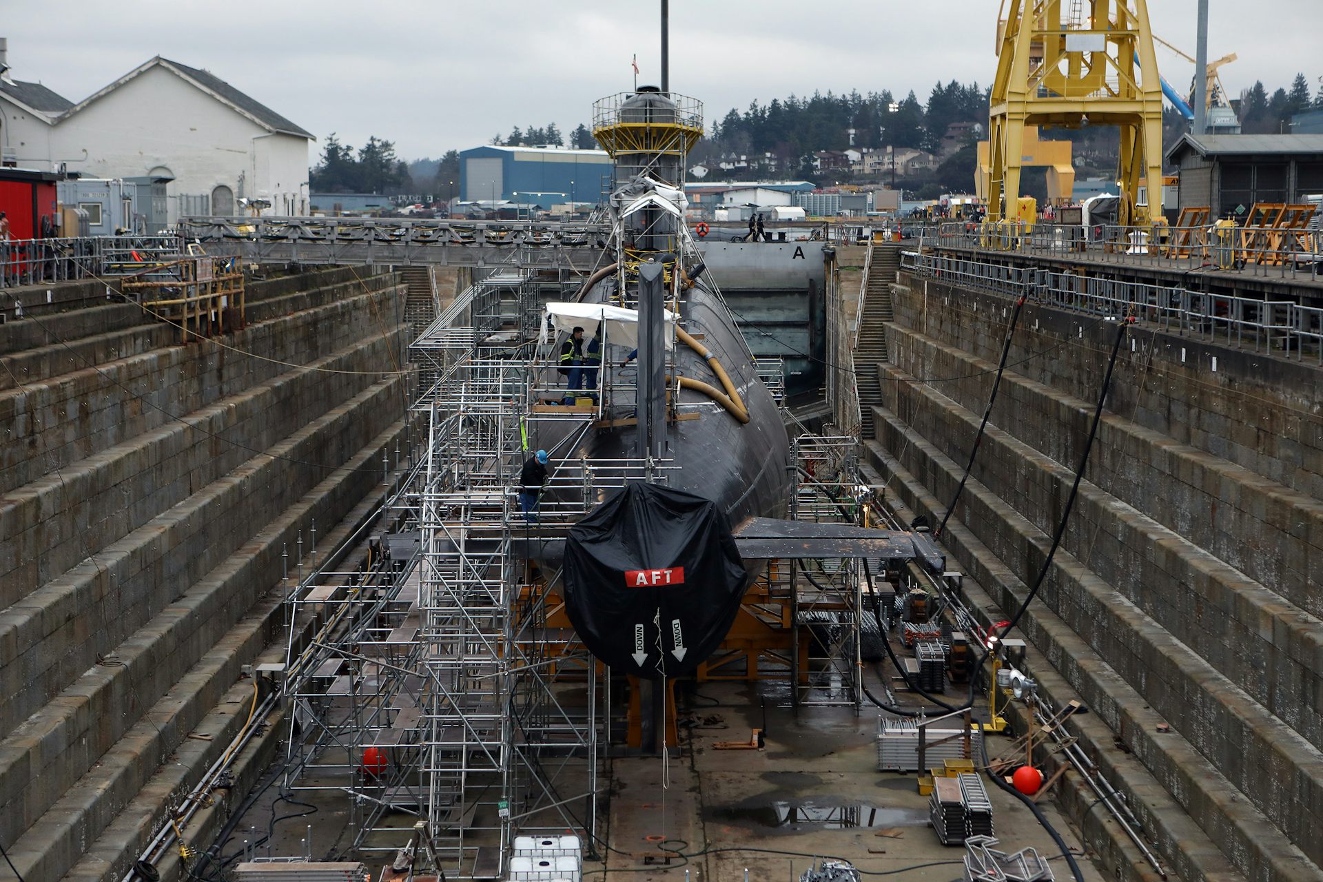 A submarine in dry dock as workers tend to it on a cloudy day.