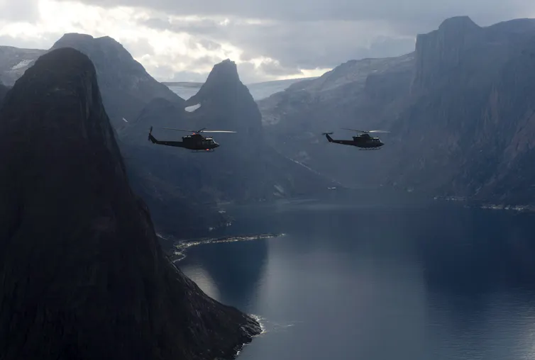 Two helicopters fly amid soaring mountains surrounding open water.